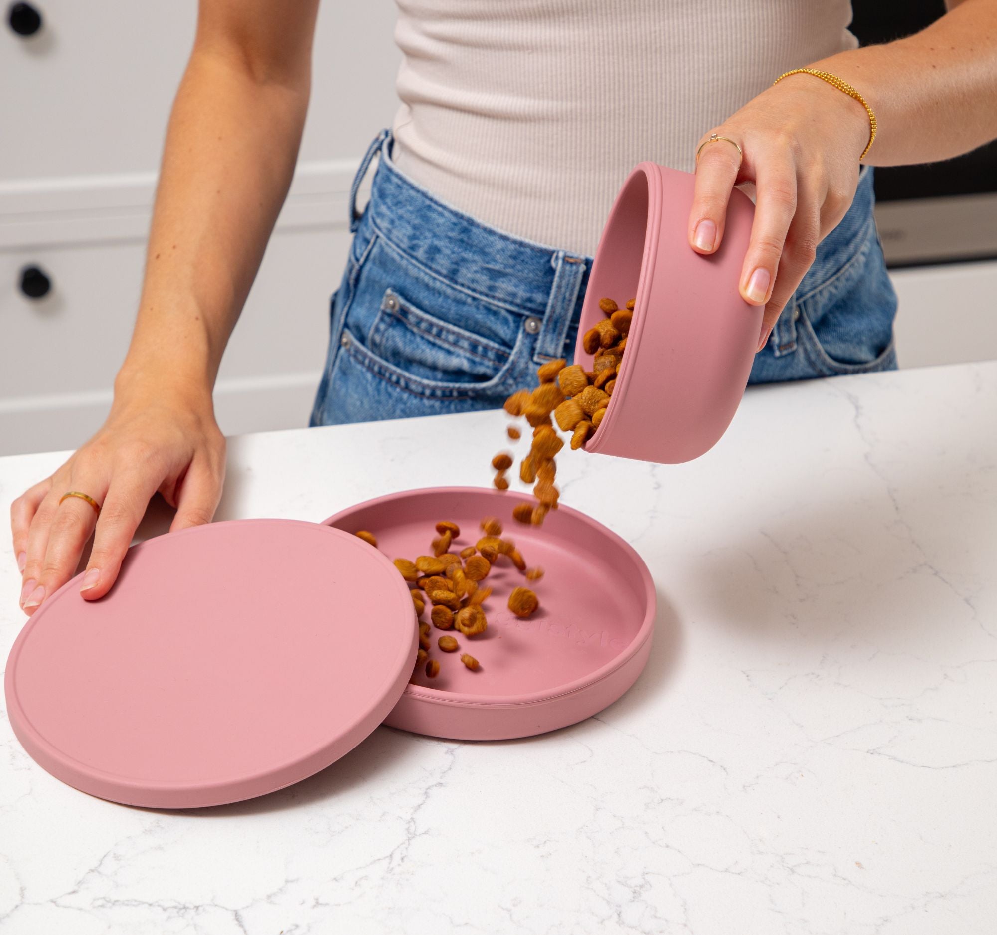 Person pouring dog food into a pink pet bowl on a white surface.